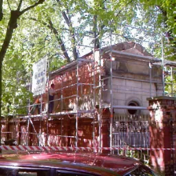 Renowacja zabytkowego grobowca z cegły, otoczonego rusztowaniami, z widocznym pracownikiem i drzewami w tle.