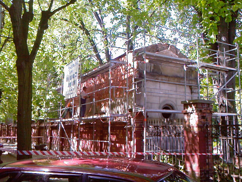 Renowacja zabytkowego grobowca z cegły, otoczonego rusztowaniami, z widocznym pracownikiem i drzewami w tle.