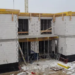 Dwukondygnacyjny dom w trakcie budowy z białych bloczków, widoczne otwory okienne i drzwiowe z drewnianym szalunkiem, częściowo ośnieżony teren budowy, w tle zabudowa miejska i dźwig.