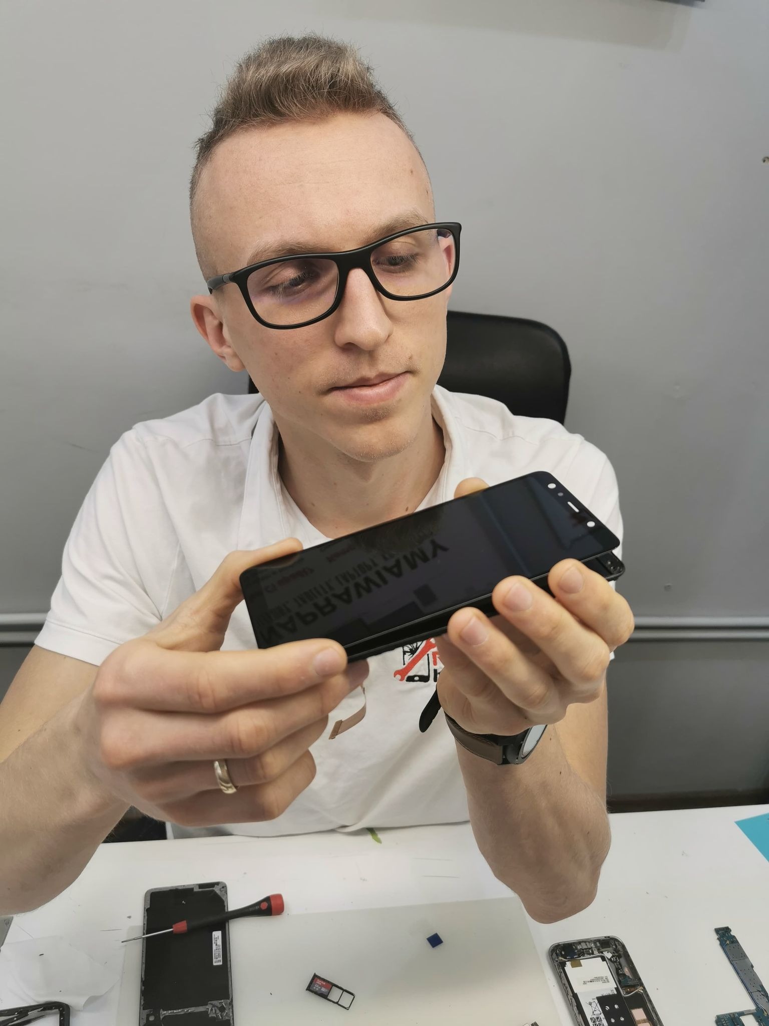 Technik z okularami trzyma w dłoniach nowy wyświetlacz do telefonu Samsung, przygotowując się do montażu. Na stole widoczne rozłożone elementy telefonu i narzędzia.