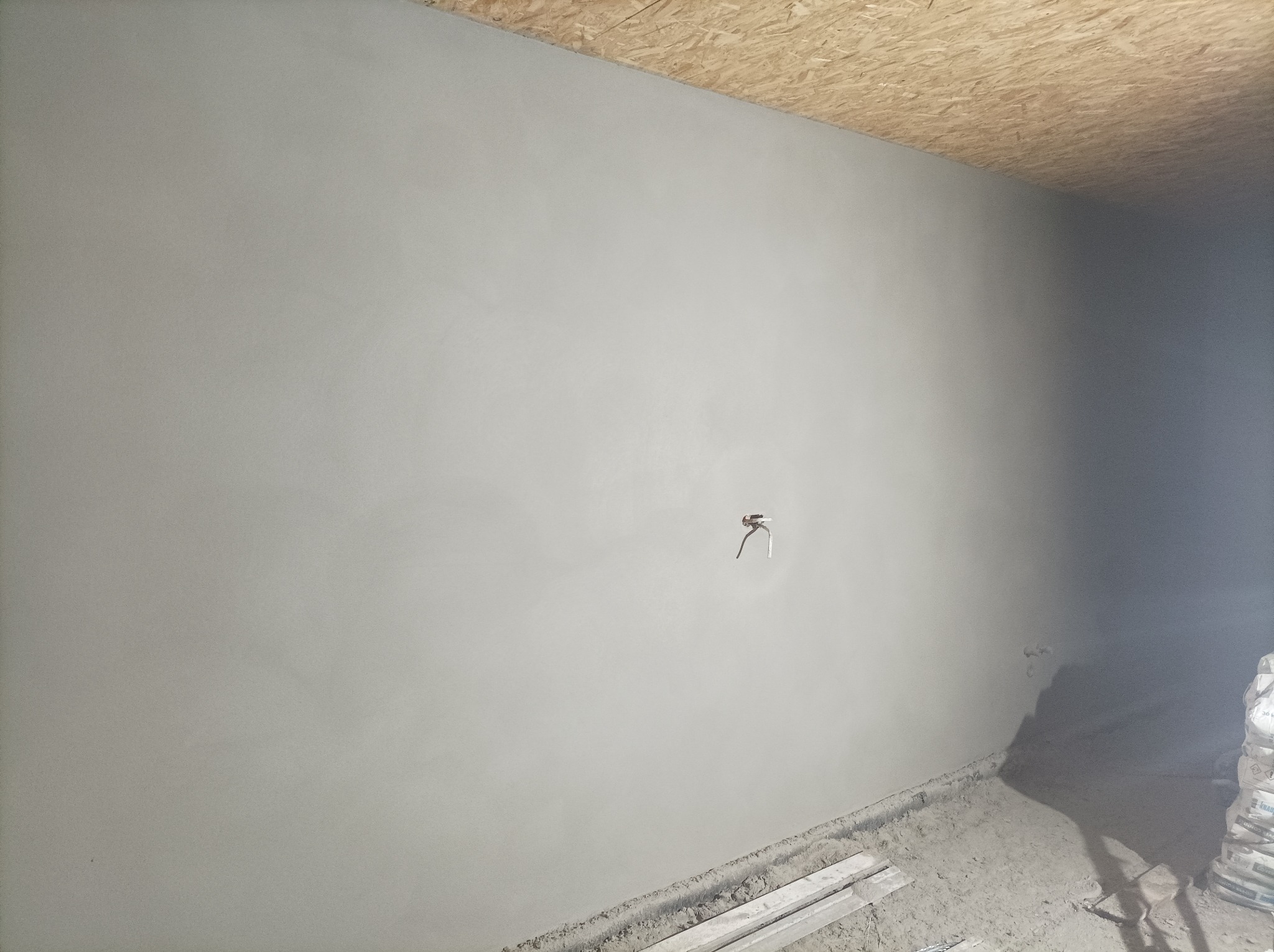 Świeżo nałożony, gładki tynk na ścianie z widocznym wystającym przewodem elektrycznym i fragmentem sufitu z płyty OSB. Na podłodze widoczne deski i worki z zaprawą.