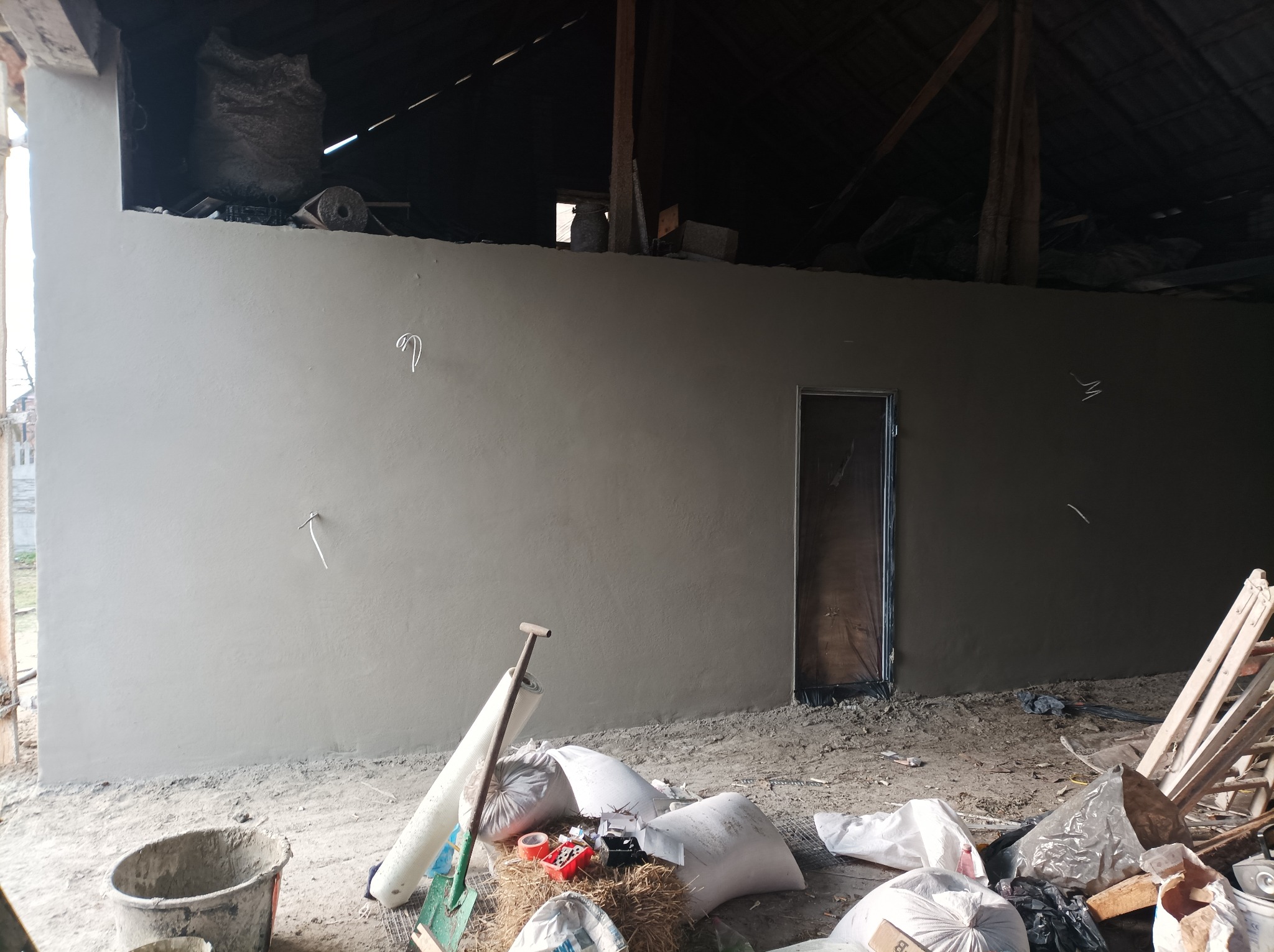 Świeżo położony, gładki, szary tynk na ścianie wewnętrznej budynku w trakcie remontu. Widoczne worki z materiałem, narzędzia budowlane i prowizoryczne oświetlenie. W ścianie osadzona surowa...