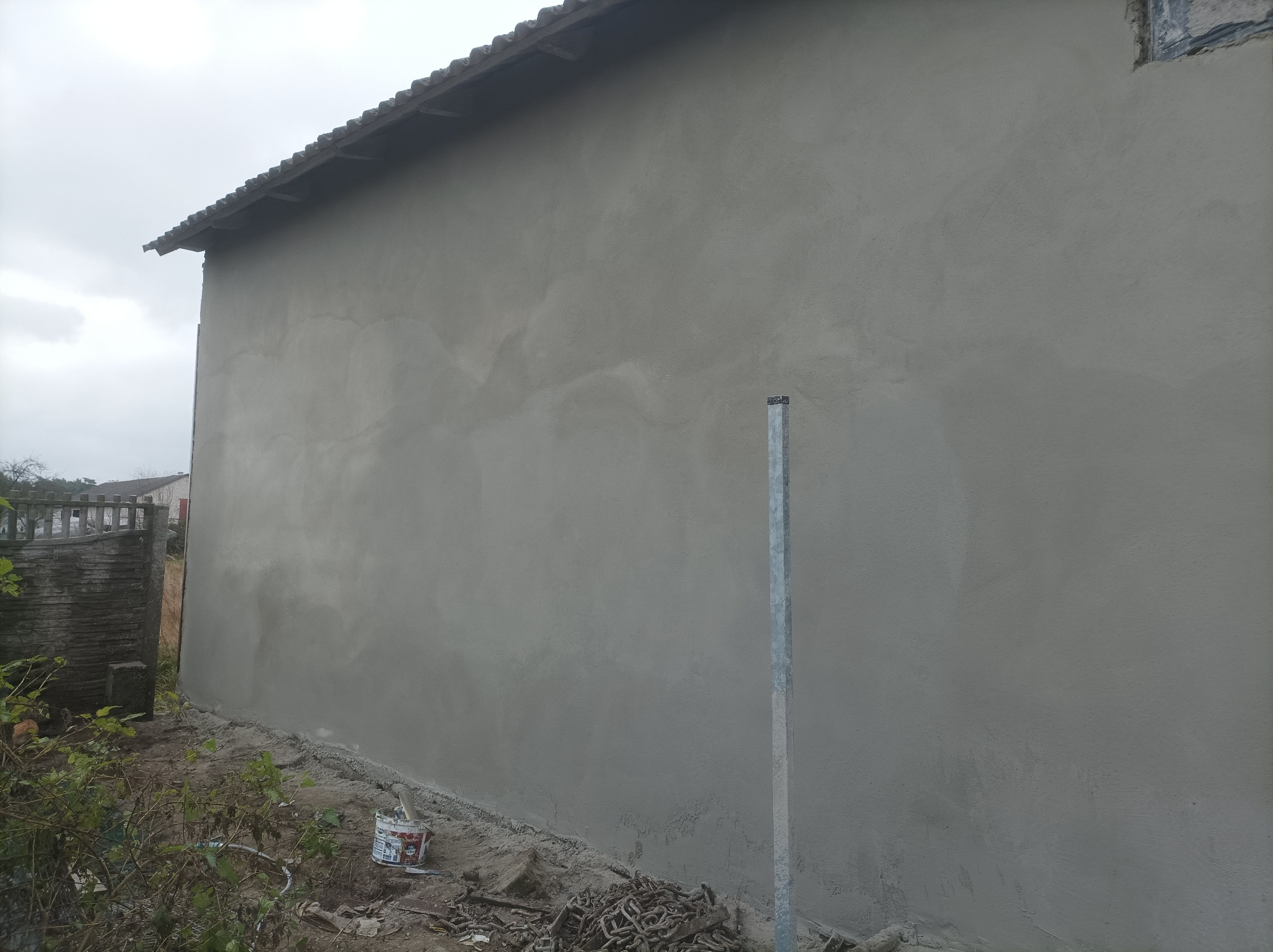 Świeżo otynkowana ściana budynku z widocznymi śladami po nałożeniu tynku, metalowy słupek i wiadro z resztkami zaprawy u podstawy ściany, teren lekko zabrudzony.