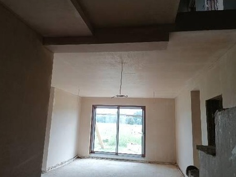 Pokój po nałożeniu tynków: surowe ściany o jasnej, jednolitej fakturze, widoczne elementy instalacji elektrycznej na suficie, duże okno z widokiem na zieleń za oknem.