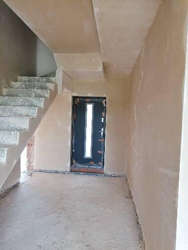 Wnętrze w trakcie prac tynkarskich: świeżo otynkowane ściany, schody z surowego betonu i czarne drzwi wejściowe z pionowym przeszkleniem.