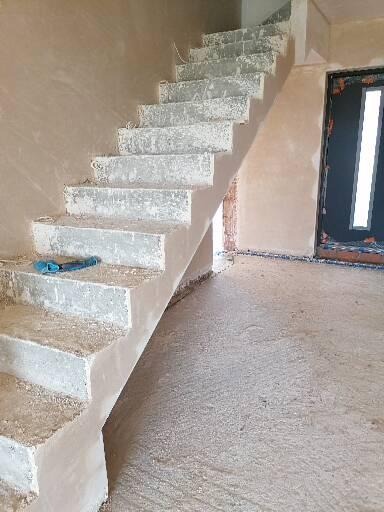 Surowe schody z nałożonym tynkiem maszynowym, widoczne niebieskie narzędzie na jednym ze stopni, ściany również pokryte tynkiem.