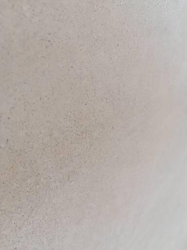 Jasnoszara, gładka powierzchnia tynku maszynowego z drobnymi, ciemnymi punktami, oświetlona z prawej strony, ukazująca fakturę i minimalne nierówności.