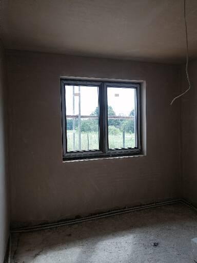 Pokój po nałożeniu gładzi gipsowej na ścianach i suficie, widoczne okno z widokiem na zieleń, szare ściany i podłoga, wystający przewód elektryczny z sufitu.