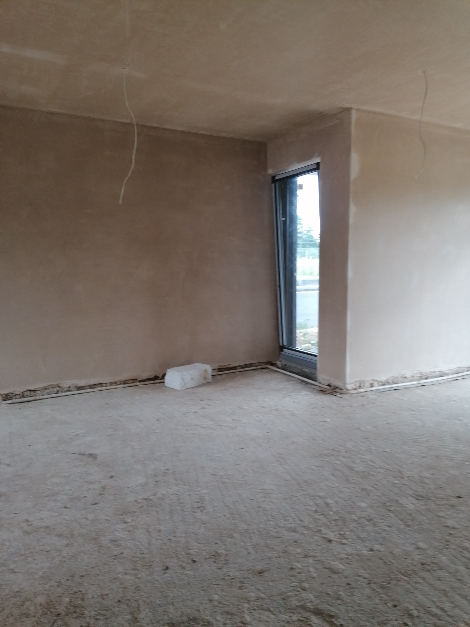 Wnętrze pomieszczenia po nałożeniu tynków: gładkie ściany w odcieniach beżu, widoczne przewody elektryczne zwisające z sufitu, okno z widokiem na zieleń, oraz biały bloczek na podłodze przy ścianie.
