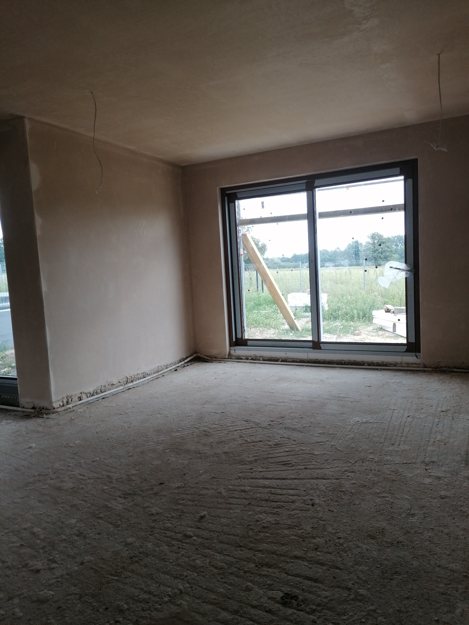 Świeżo otynkowane ściany w jasnym pomieszczeniu z dużym oknem przesuwnym, widoczne niedoskonałości na podłodze i wystające kable z sufitu.