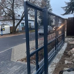 POL.ART-STAL VADYM POLISHCHUK - Szara, przesuwna brama z paneli z widocznym systemem jezdnym, zamontowana na posesji z kostką brukową, widok z boku na tle paczkomatu i zabudowań.
