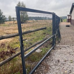 POL.ART-STAL VADYM POLISHCHUK - Otwarta brama panelowa z szarej siatki na posesji z drewnianym płotem i kamienistym podłożem, widok na pole w tle.