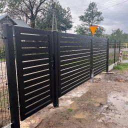 POL.ART-STAL VADYM POLISHCHUK - Czarna, nowoczesna brama wjazdowa z poziomymi szczeblami, częściowo otwarta, widoczna w błotnistym terenie na tle drzew i znaku drogowego.