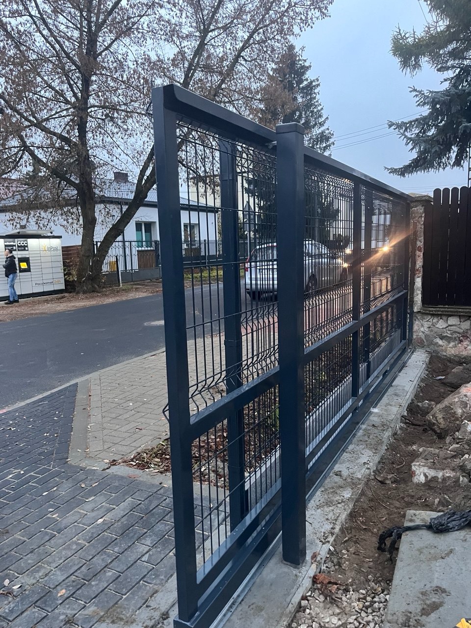 Szara, przesuwna brama z paneli z widocznym systemem jezdnym, zamontowana na posesji z kostką brukową, widok z boku na tle paczkomatu i zabudowań.