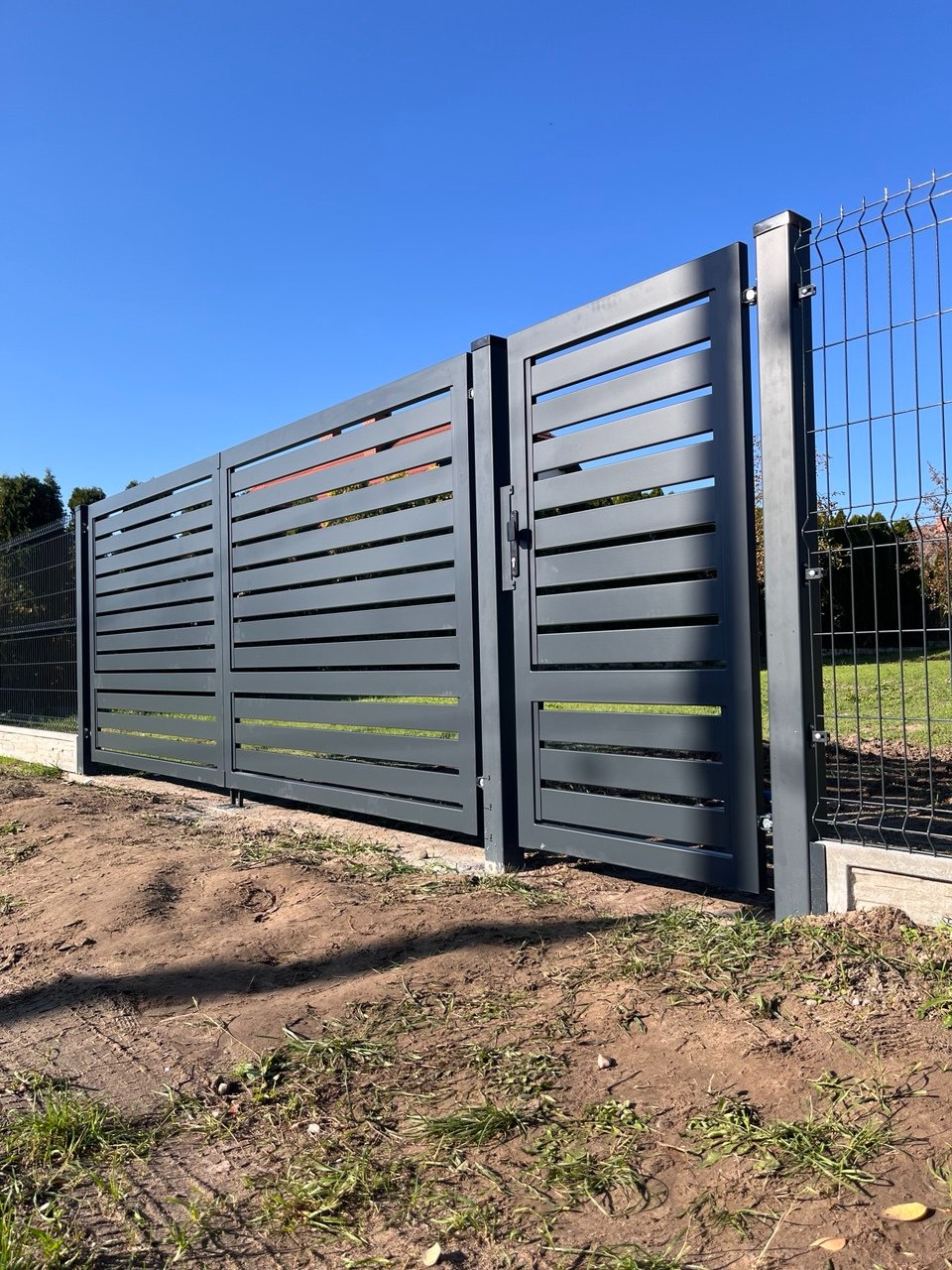 Nowoczesna brama przesuwna z poziomymi panelami w kolorze grafitowym, z widoczną furtką i fragmentem ogrodzenia z siatki, na tle błękitnego nieba.