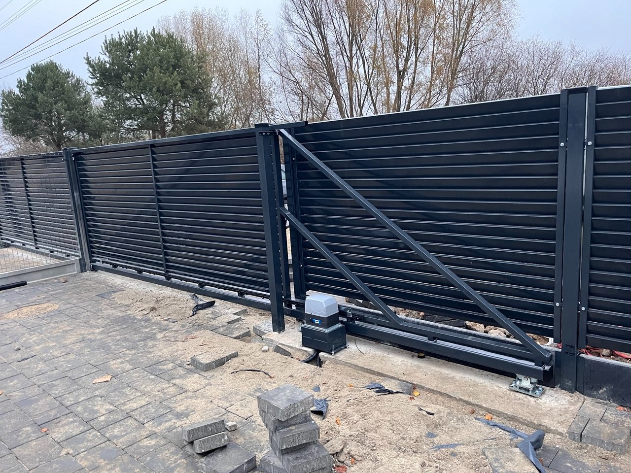 Nowoczesna, przesuwna brama wjazdowa z czarnymi, poziomymi panelami, widoczny mechanizm automatycznego otwierania i przygotowane podłoże z kostki brukowej.