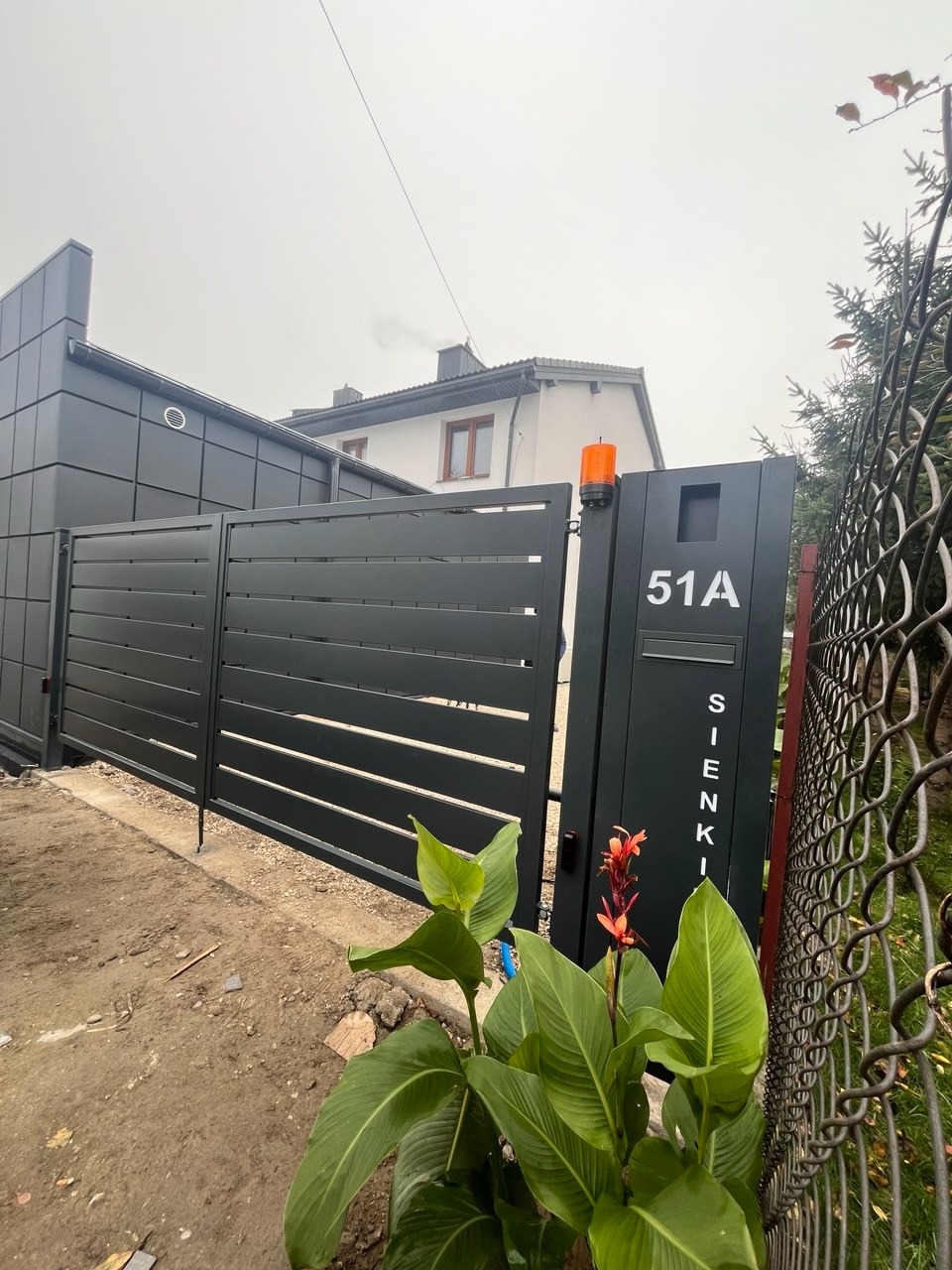 Nowoczesna, pozioma brama wjazdowa z numerem domu 51A, pomarańczowym światłem ostrzegawczym i rośliną w pierwszym planie, obok fragment ogrodzenia z siatki.