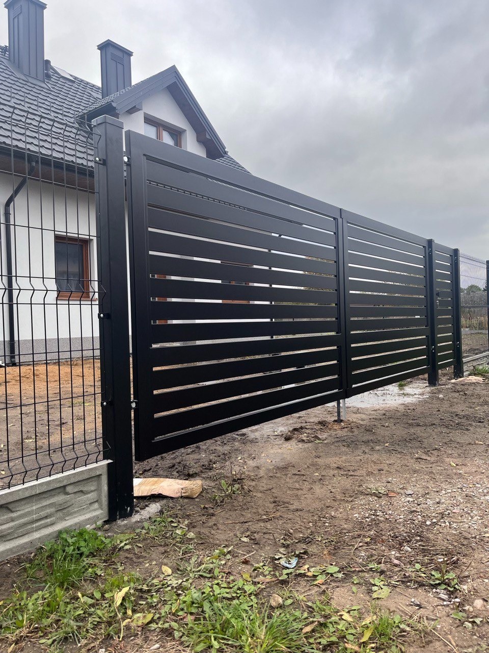 Nowoczesne, czarne ogrodzenie panelowe z poziomymi szczeblami, częściowo otwarte, z widocznym fragmentem domu w tle i siatką ogrodzeniową po prawej stronie.