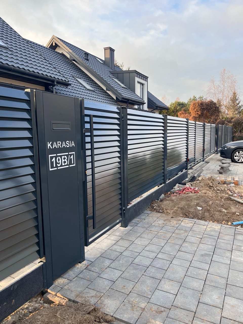 Nowoczesne, poziome ogrodzenie lamelowe w kolorze grafitowym z bramką i numerem domu 19B/1, widoczne kostka brukowa i fragment domu z ciemnym dachem