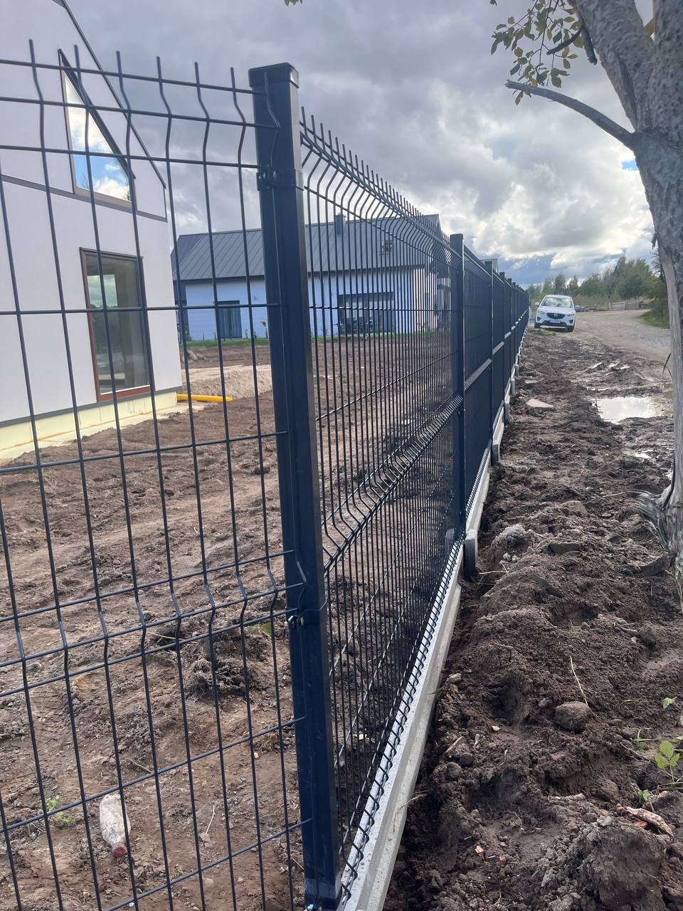 Ciemne panele ogrodzeniowe z betonową podmurówką, widok z boku, w tle domy i samochód na błotnistej drodze.