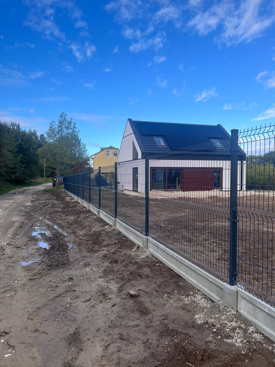 Nowoczesny dom z ciemną dachówką otoczony grafitowym ogrodzeniem panelowym na betonowej podmurówce, Grabce Józefpolskie. Teren wokół ogrodzenia częściowo błotnisty.