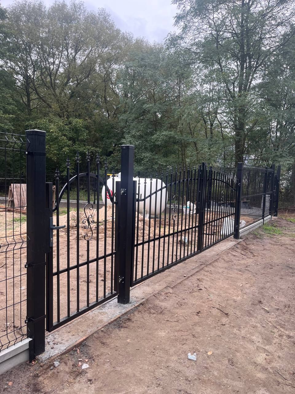 Czarna brama wjazdowa z elementami dekoracyjnymi i przylegającym ogrodzeniem panelowym z siatki, widoczny zbiornik na gaz w tle, teren wyrównany, bez trawy.
