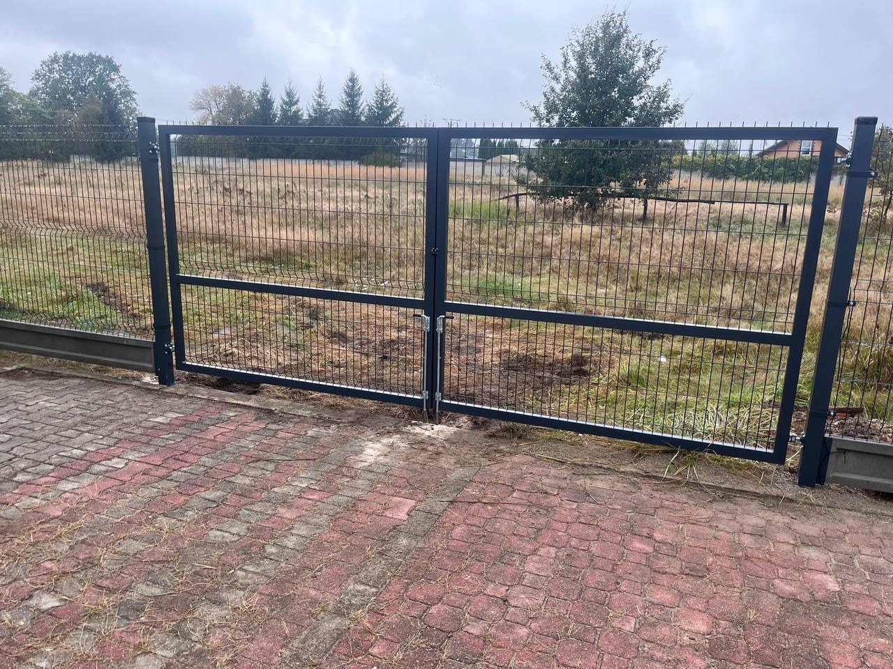 Dwuskrzydłowa, metalowa brama wjazdowa z wypełnieniem z siatki panelowej, zamontowana na posesji z kostką brukową, widok na pole z trawą i drzewami w tle.