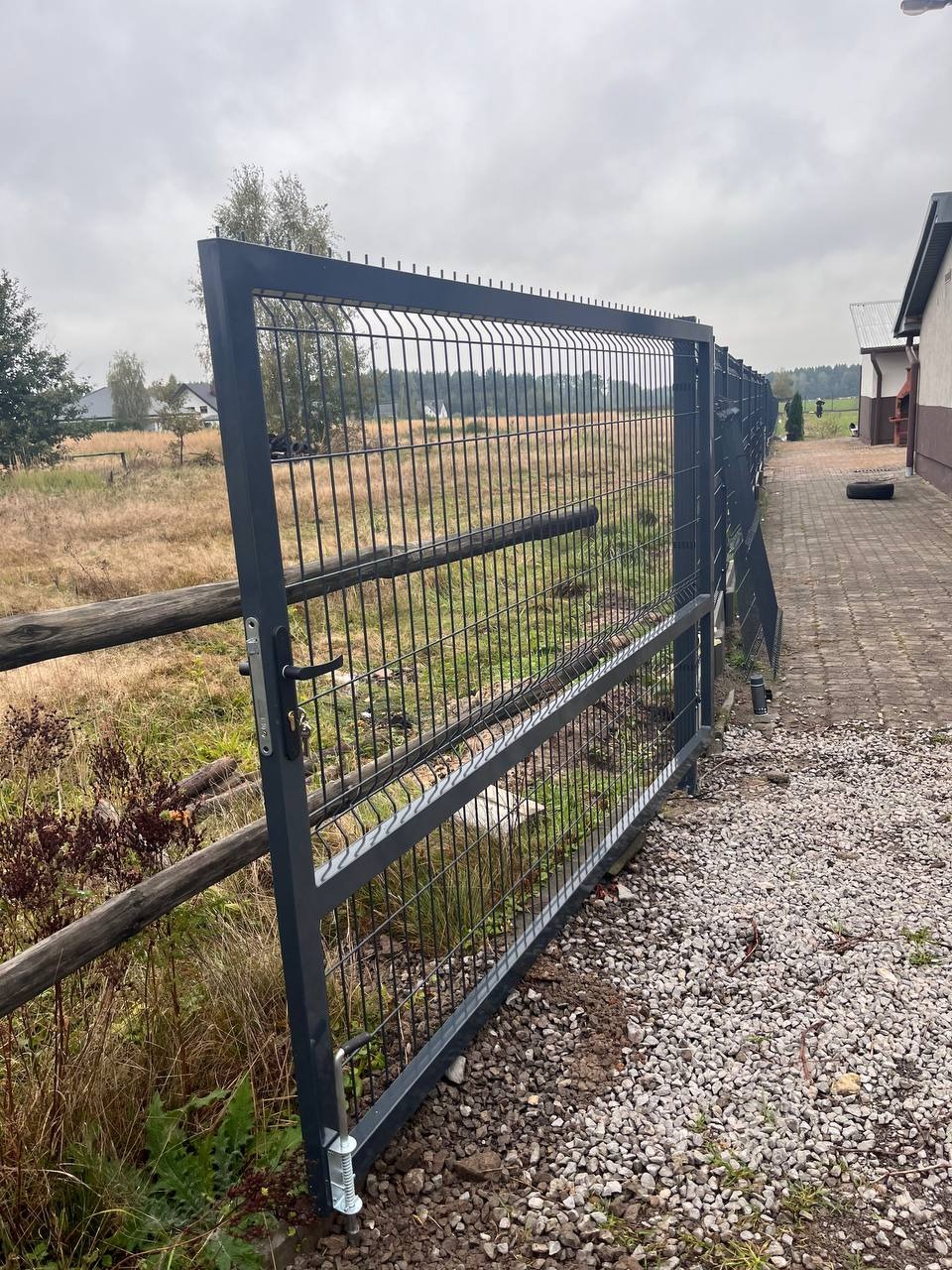 Otwarta brama panelowa z szarej siatki na posesji z drewnianym płotem i kamienistym podłożem, widok na pole w tle.