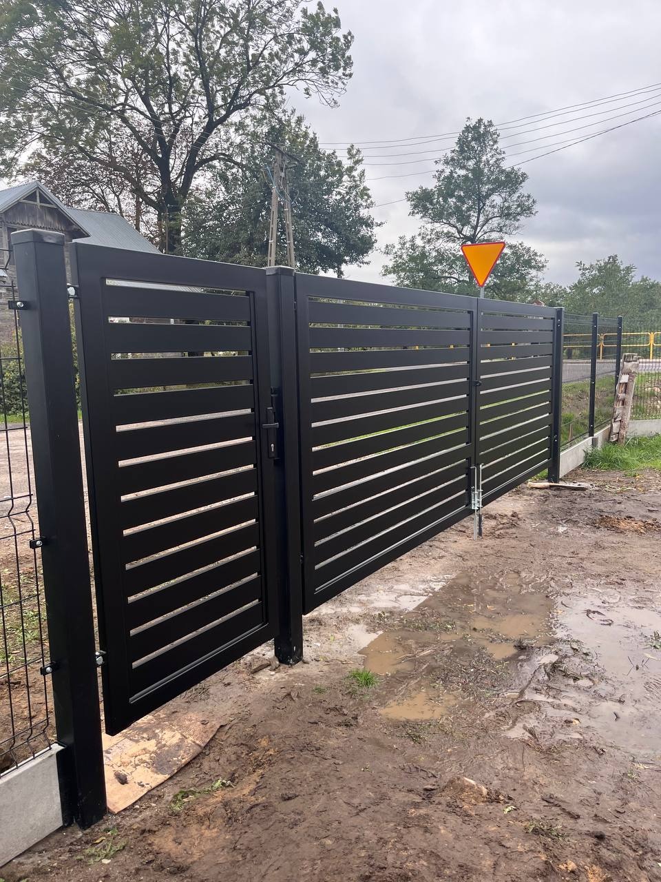 Czarna, nowoczesna brama wjazdowa z poziomymi szczeblami, częściowo otwarta, widoczna w błotnistym terenie na tle drzew i znaku drogowego.