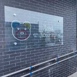 Szklana tablica z logo żłobka 'Żłobek w Kostrzynie', przymocowana do ściany z ciemnej cegły, widoczne poręcze dla niepełnosprawnych z niebieskimi zabezpieczeniami.