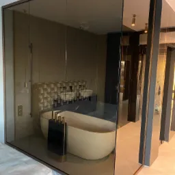 Szklana kabina prysznicowa z widoczną wolnostojącą wanną w luksusowej łazience z beżowymi płytkami i złotymi akcentami, widok z zewnątrz na zapakowany materac.