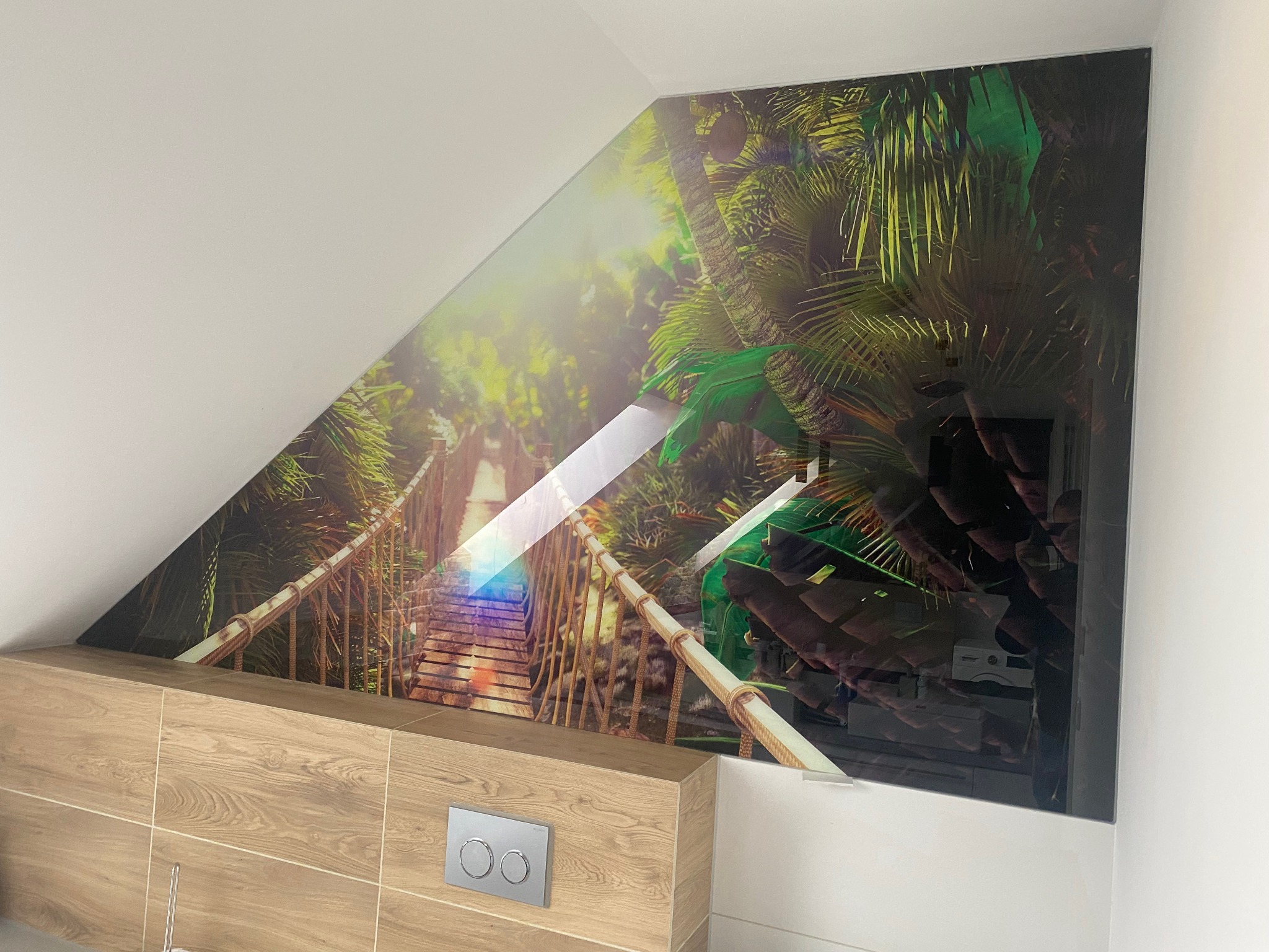 Szklany panel ścienny z tropikalnym motywem mostu linowego w łazience na poddaszu, zamontowany nad zabudową WC.