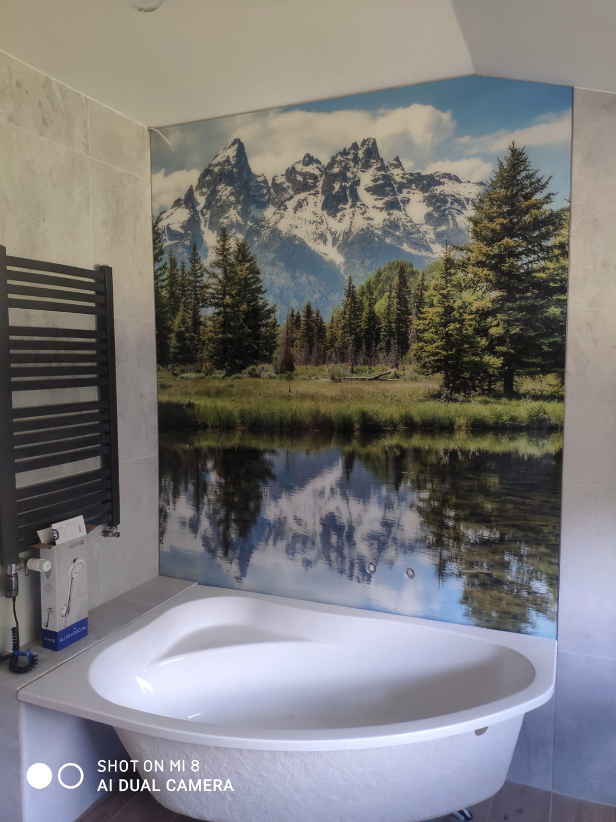 Biała narożna wanna pod ścianą z fototapetą przedstawiającą górski krajobraz z jeziorem, obok grzejnik drabinkowy.