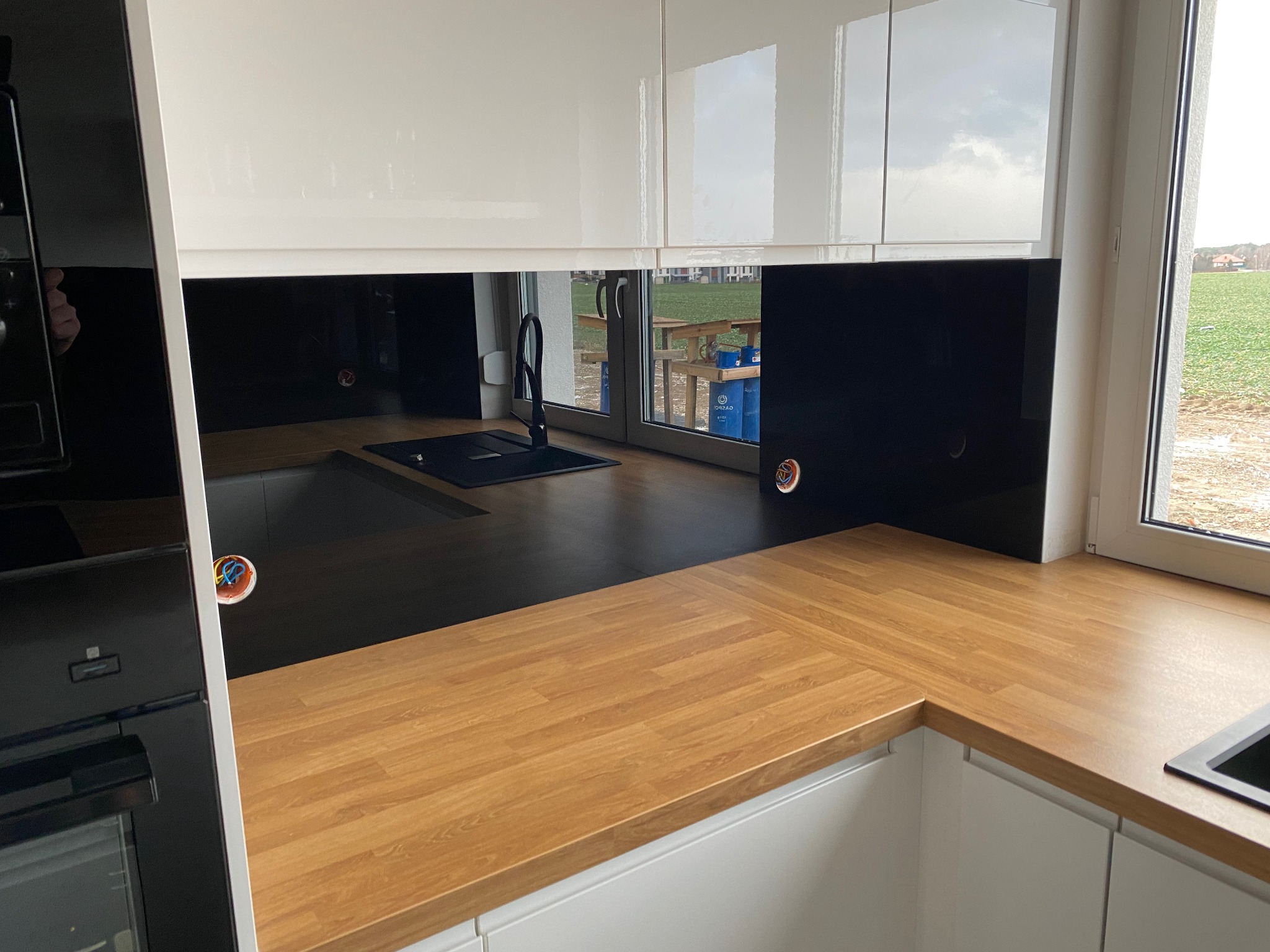 Nowoczesna kuchnia z czarnym szklanym panelem nad blatem z drewnianym wzorem, widoczne gniazdka elektryczne i zlewozmywak z czarną armaturą.