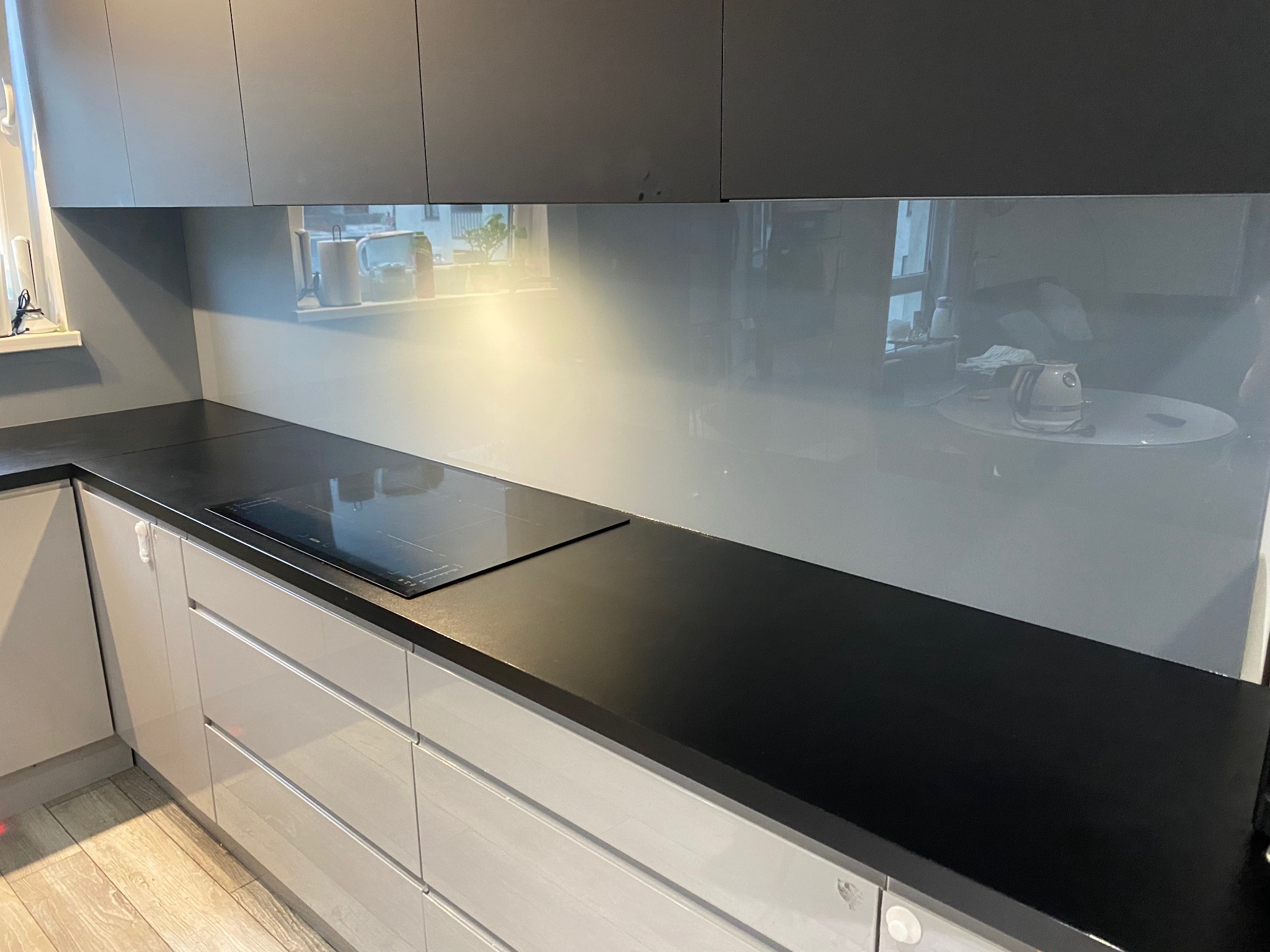 Szklany panel ścienny w kuchni, zamontowany nad czarnym blatem i płytą indukcyjną, odbijający światło i widok z okna.