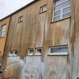 Mycie zabrudzonej elewacji budynku z wysokości podnośnika koszowego, widoczne efekty czyszczenia strumieniem wody pod ciśnieniem.