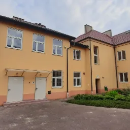 Dwupiętrowy budynek z żółtą elewacją, czerwoną dachówką i kostką brukową na dziedzińcu, widok z dołu na pochmurnym niebie.