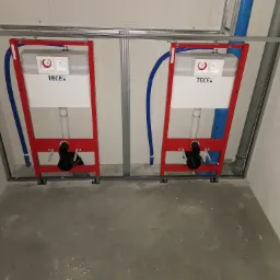 Dwa podtynkowe stelaże WC marki TECE w czerwonej ramie, zamontowane w konstrukcji z profili stalowych, z widocznymi niebieskimi rurami doprowadzającymi wodę, na surowym betonowym podłożu w trakcie...