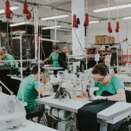 Szeroki plan hali produkcyjnej z rzędem stanowisk krawieckich, gdzie kobiety w zielonych koszulkach szyją na maszynach, a nad nimi wiszą czerwone gniazda zasilające.