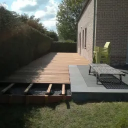 Drewniany taras w trakcie budowy, widoczne deski, podkonstrukcja i fragment wykończonej części z leżakiem na tle ceglanego domu i żywopłotu.