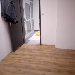 PUH Stolkam - Widok na świeżo położoną podłogę panelową w korytarzu, z widoczną częścią drzwi wejściowych z kurtką zawieszoną na klamce i białymi drzwiami na końcu korytarza.