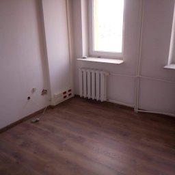 PUH Stolkam - Puste pomieszczenie z dwoma oknami, grzejnikiem i panelami podłogowymi w odcieniach brązu, widoczne niedokończone prace elektryczne przy ścianie.