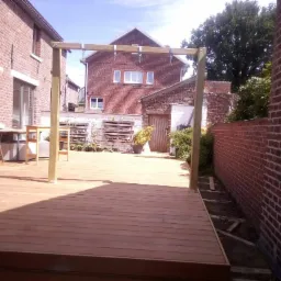 Nowo wybudowany drewniany taras z pergolą w ogrodzie, otoczony ceglanymi budynkami i zielenią.