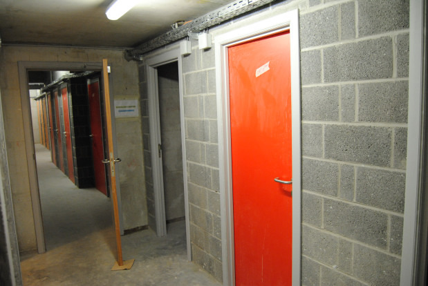 Długi korytarz piwniczny z rzędem boksów magazynowych oddzielonych ścianami z betonowych pustaków, zamykanych na metalowe drzwi; jeden boks ma drzwi w kolorze intensywnej czerwieni.
