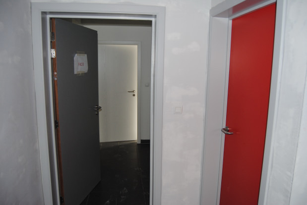 Wnętrze z dwoma nowo zamontowanymi drzwiami: szare skrzydło z naklejką F408 i czerwone skrzydło z klamką ze stali nierdzewnej, widoczne ślady po malowaniu na ścianach.