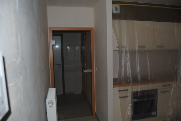 Wnętrze w trakcie remontu: widoczna kuchnia osłonięta folią malarską, surowa ściana z grzejnikiem i otwarte drzwi do łazienki z metalową zasuwką.
