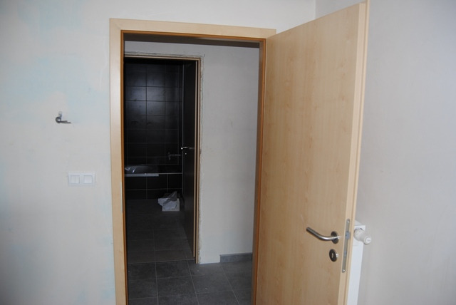 Beżowe, otwarte drzwi wewnętrzne z widocznym fragmentem łazienki z czarnymi płytkami w tle, po lewej stronie ściana z włącznikiem i hakiem.