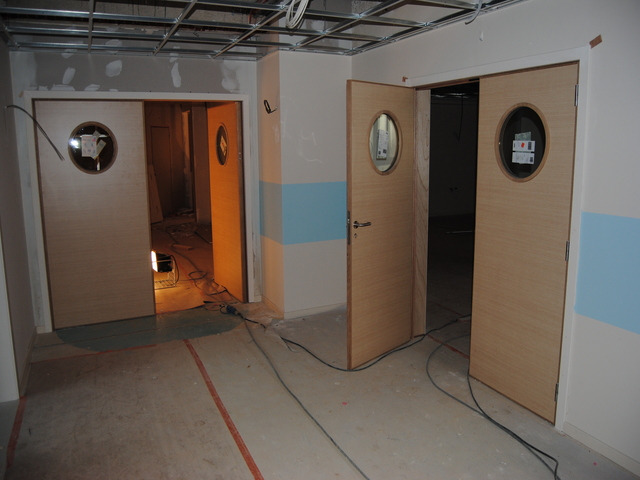 Dwa komplety drewnopodobnych drzwi z okrągłymi okienkami, w trakcie prac wykończeniowych w pomieszczeniu; widoczne przewody, lampa budowlana i niedokończony sufit podwieszany.