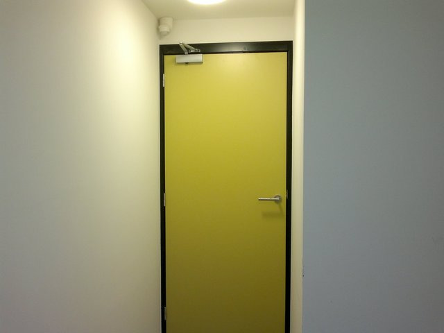 Wewnętrzne drzwi w kolorze jaskrawej zieleni z czarną ramą, wyposażone w klamkę i mechanizm samozamykający, zamontowane w białej ścianie korytarza.