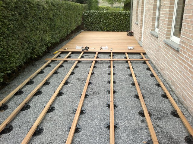 Konstrukcja drewnianego tarasu w trakcie budowy, legary na regulowanych wspornikach na żwirowym podłożu, częściowo ułożona deska tarasowa przy ścianie budynku.